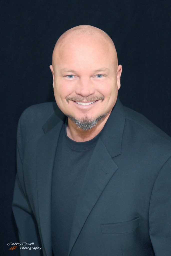 Smiling bald man in black suit against dark background.